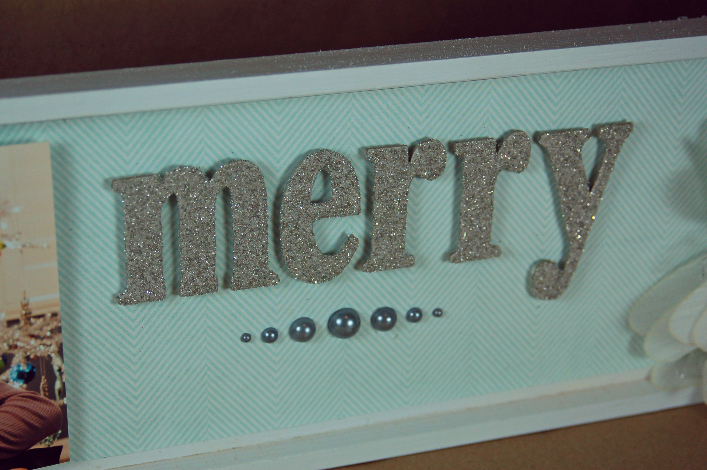 close up of chipboard letters that spell out merry and are covered in silver glass glitter against a soft, light blue patterned paper