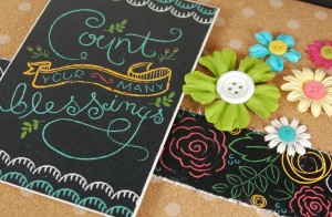 close up of black shadow box with cork background and chalkboard-style paper. There are flower and button accents, and the message says count your blessings