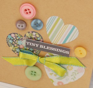 Cover of kraft album with hearts, a bow, and buttons in different light colors with a sticker that says tiny blessings
